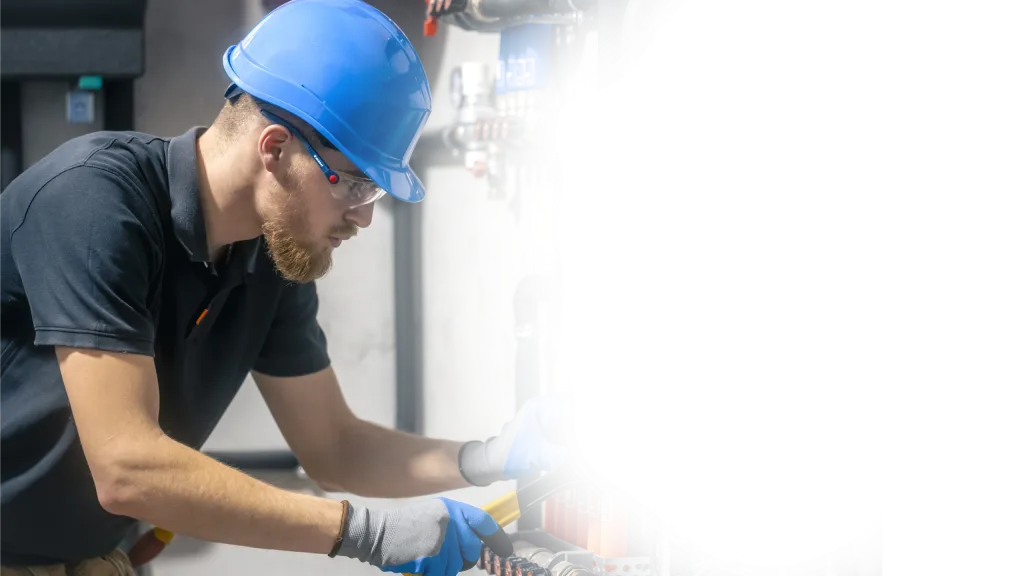Installateur mit blauem Schutzhelm und Schutzbrille arbeitet an Rohrleitungen in einem Technikraum