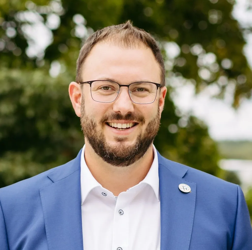 Portraetfoto von Kraus Julian im blauen Sakko, weißem Hemd, Brille und Bart